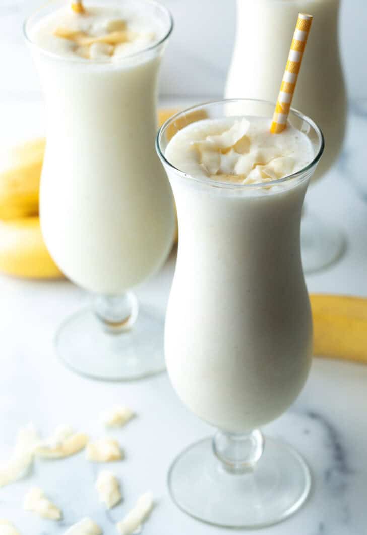 3 large mix drink glasses with banana smoothie and a yellow striped straw in each glass.