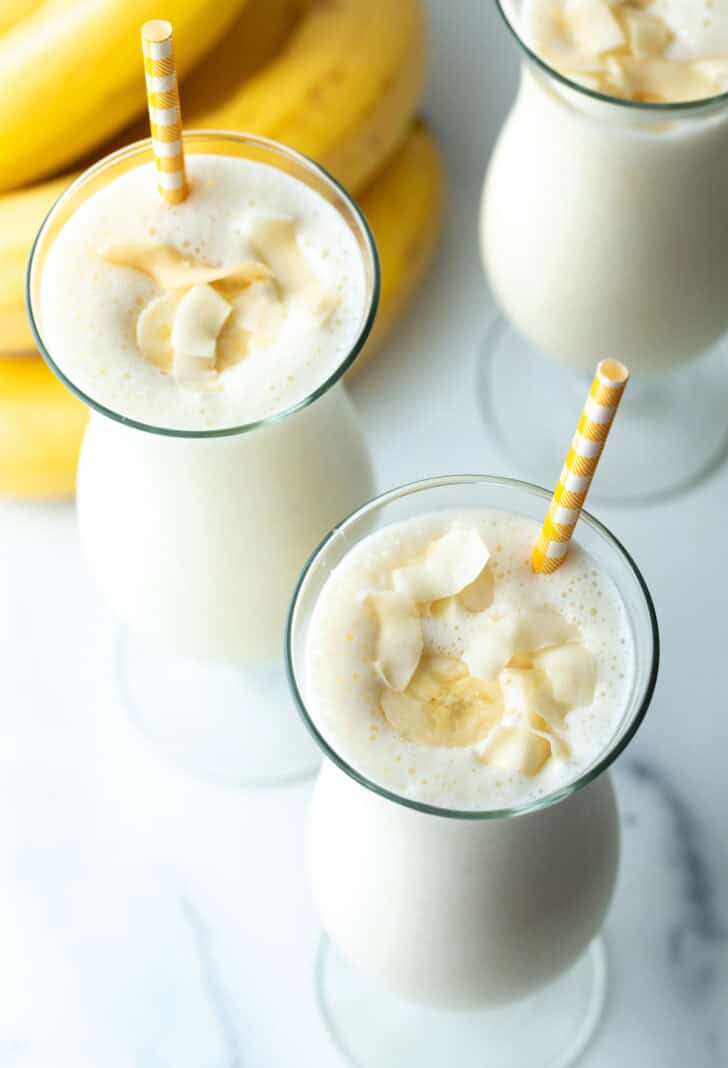 Top down view of a large mixed drink glass with banana smoothie and a yellow striped straw in each glass.