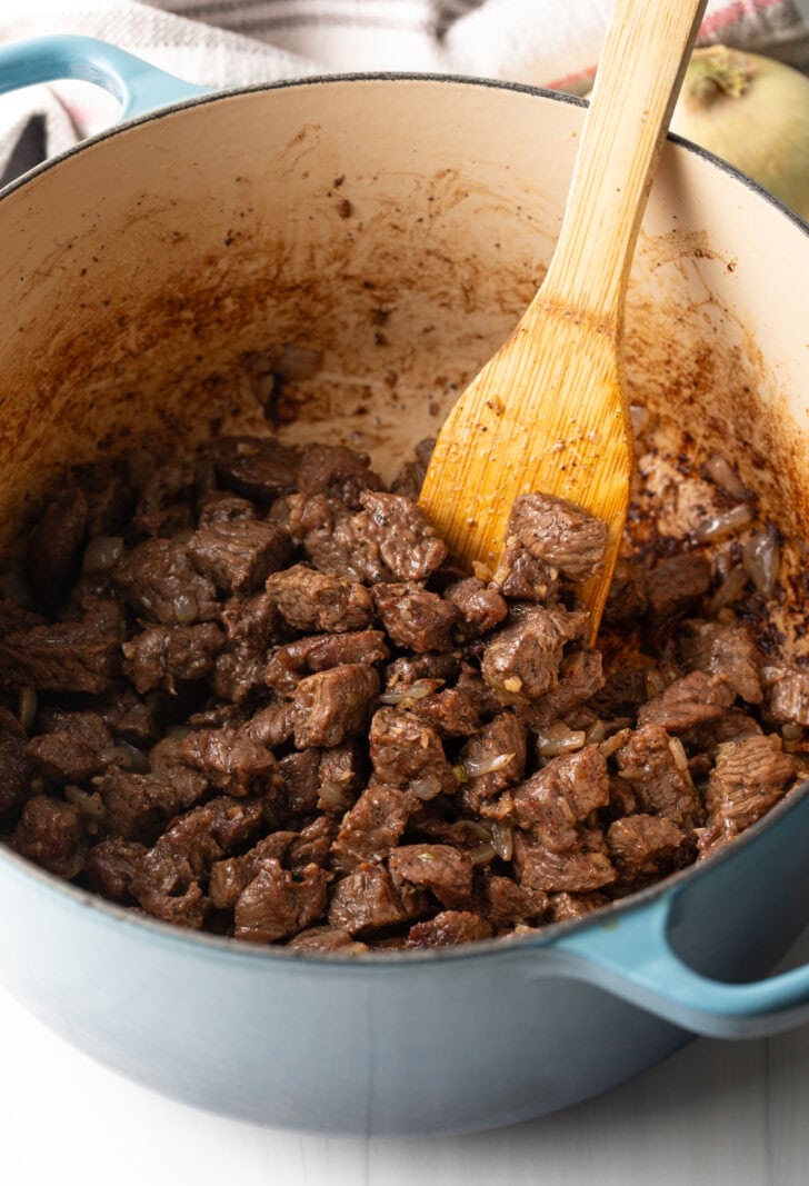 Wood spatula stirring pieces of beef as they cook in a large pot.