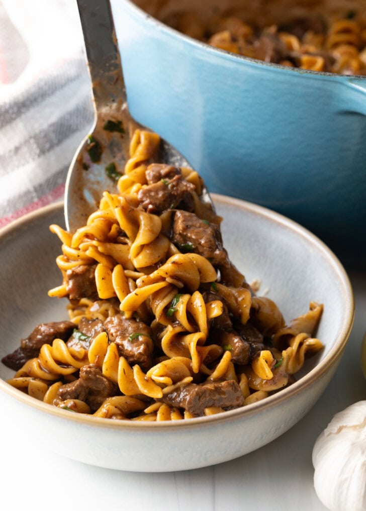 Metal spoon dishing out a serving of noodles with beef into a white bowl.