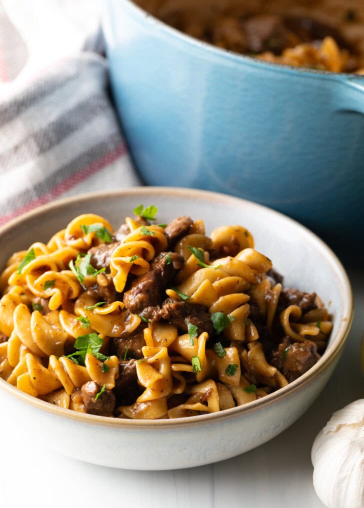 White bowl with serving of beef and noodles, topped with fresh chopped parsley.