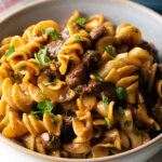 White bowl with serving of beef and noodles, topped with fresh chopped parsley.