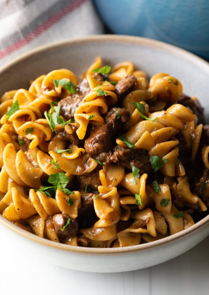 White bowl with serving of beef and noodles, topped with fresh chopped parsley.