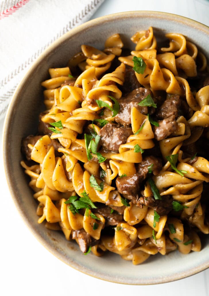 Close view white bowl with serving of beef and noodles, topped with fresh chopped parsley.