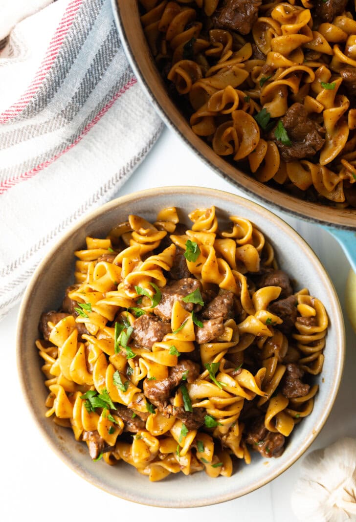 White bowl with serving of beef and noodles, topped with fresh chopped parsley.