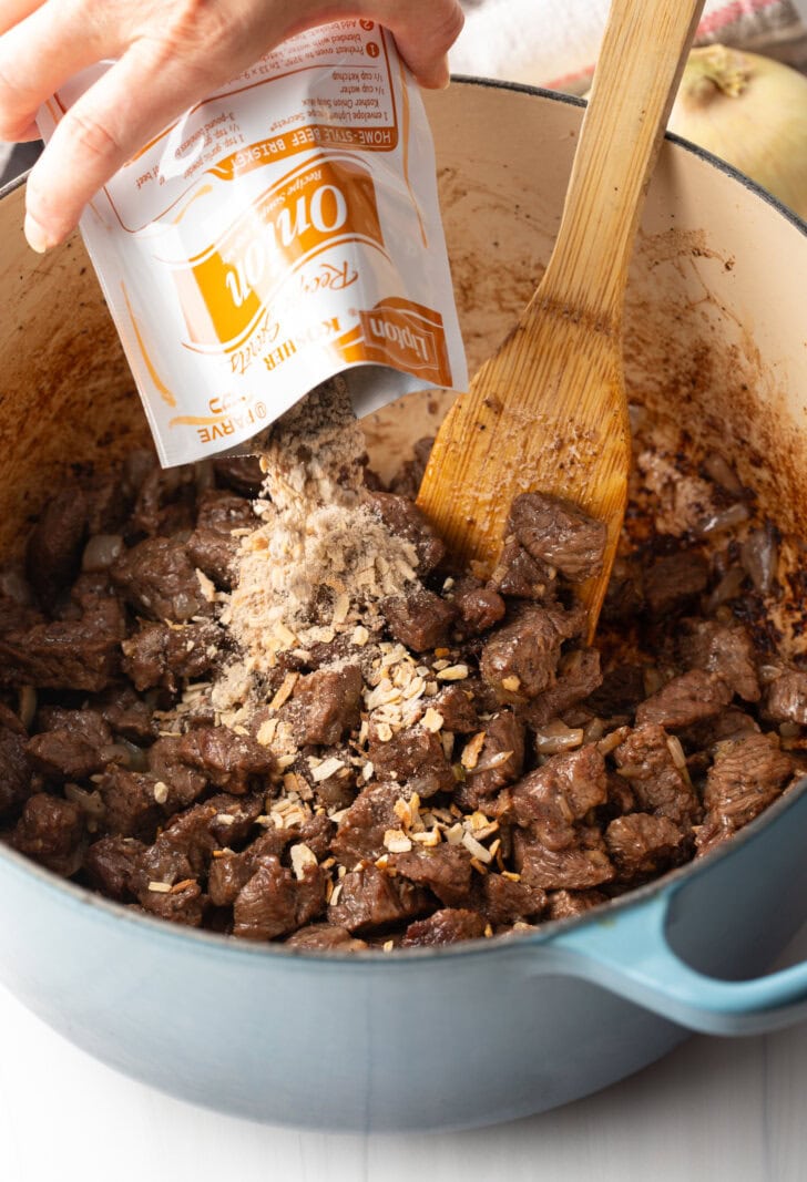 Adding seasonings from a onion soup pack to the cooked beef in a large pot, with wood spatula.