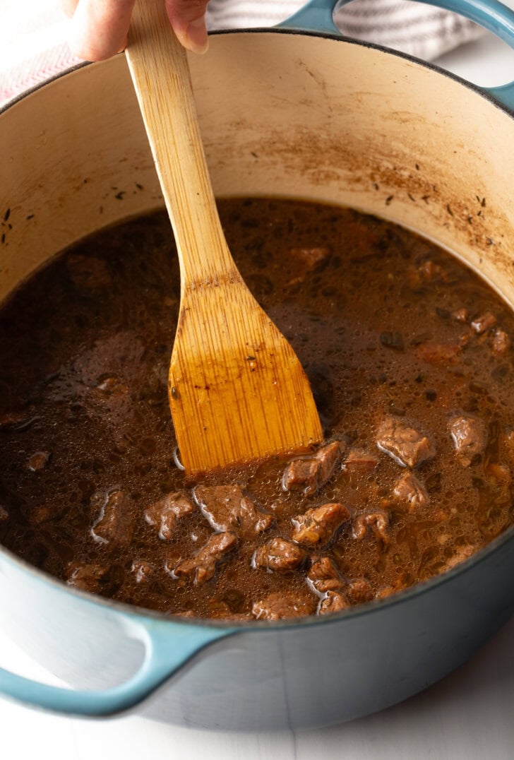 Wooden spoon stirring pieces of cooked beef in gravy in a large cooking pot.