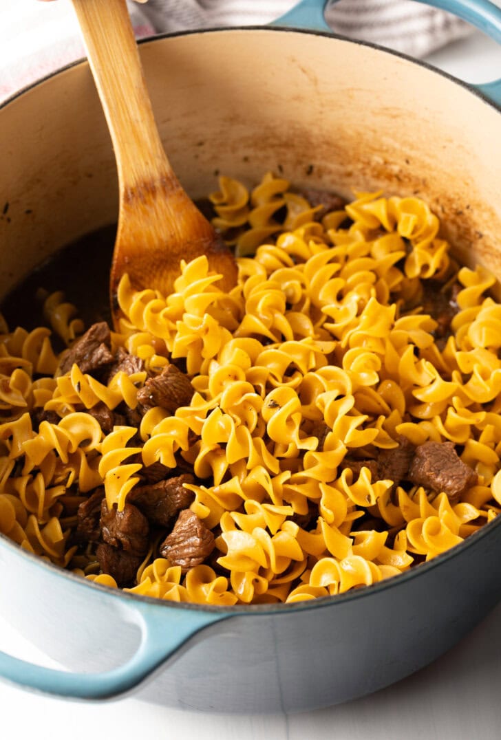 Wood spatula stirring egg noodles into the cooking pot with pieces of beef.