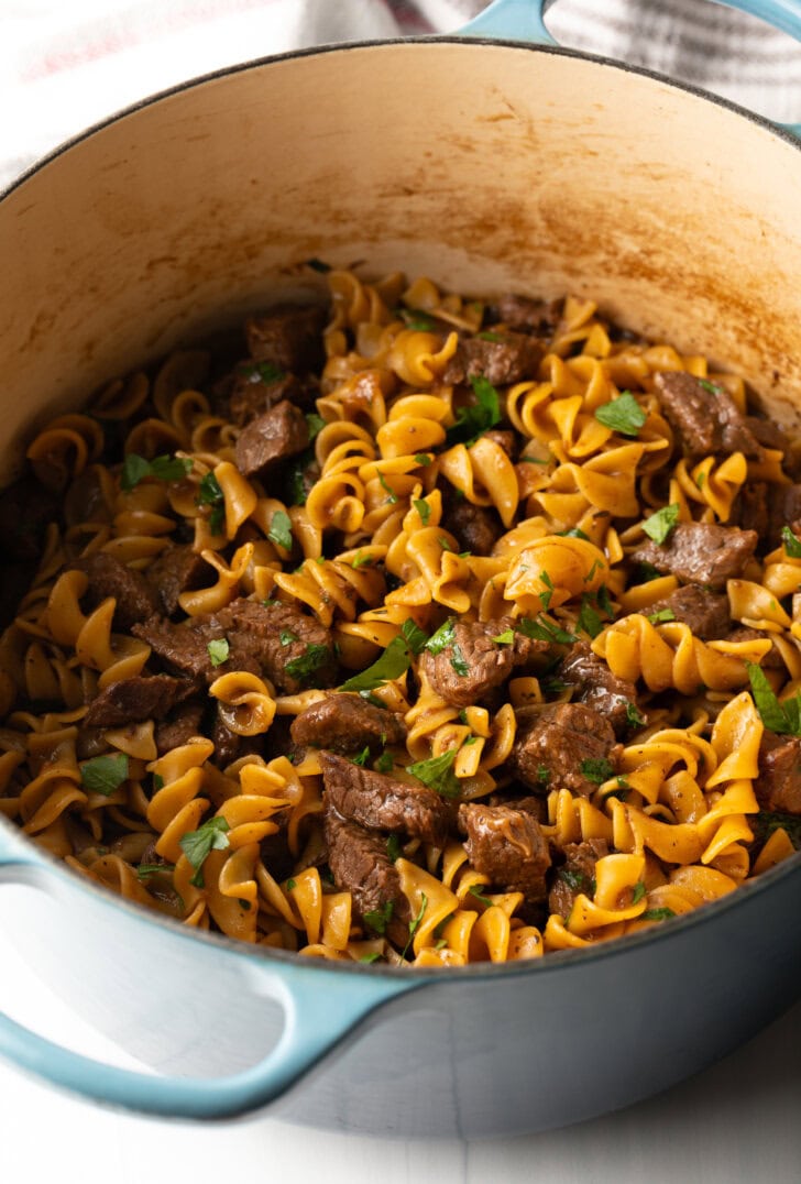 Top down view egg noodles and pieces of cooked beef in a large pot. Topped with sprinkling of fresh chopped parsley.