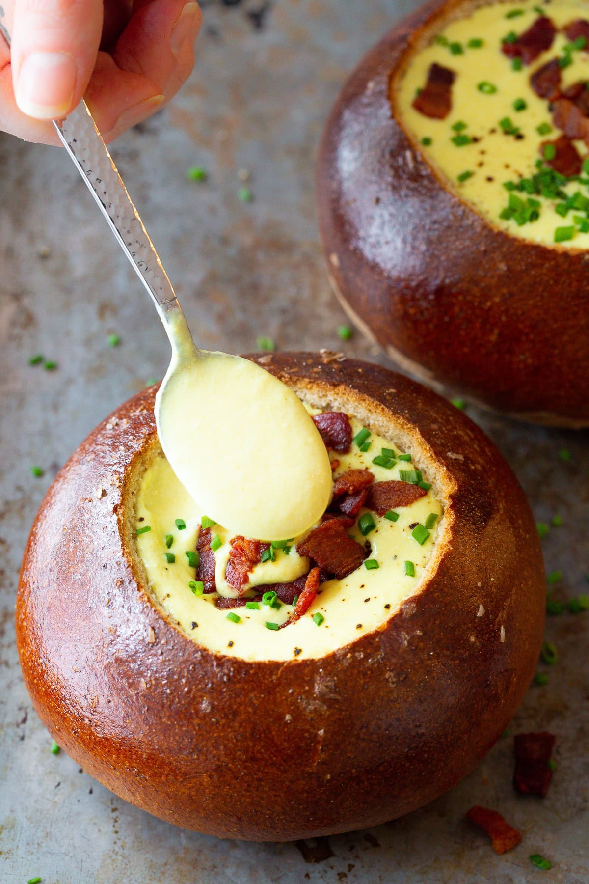 Spoon with cheese soup in it being held above a bread bowl filled with soup. 