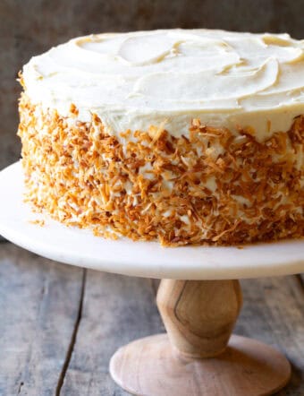 Three layer carrot cake covered in white frosting and toasted coconut. The cake is on a white pedestal cake stand.