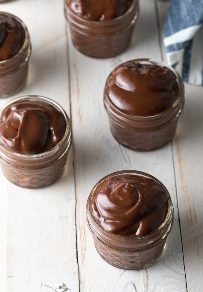 Small glass jars lined up on a wooden background and filled with chocolate pudding. 