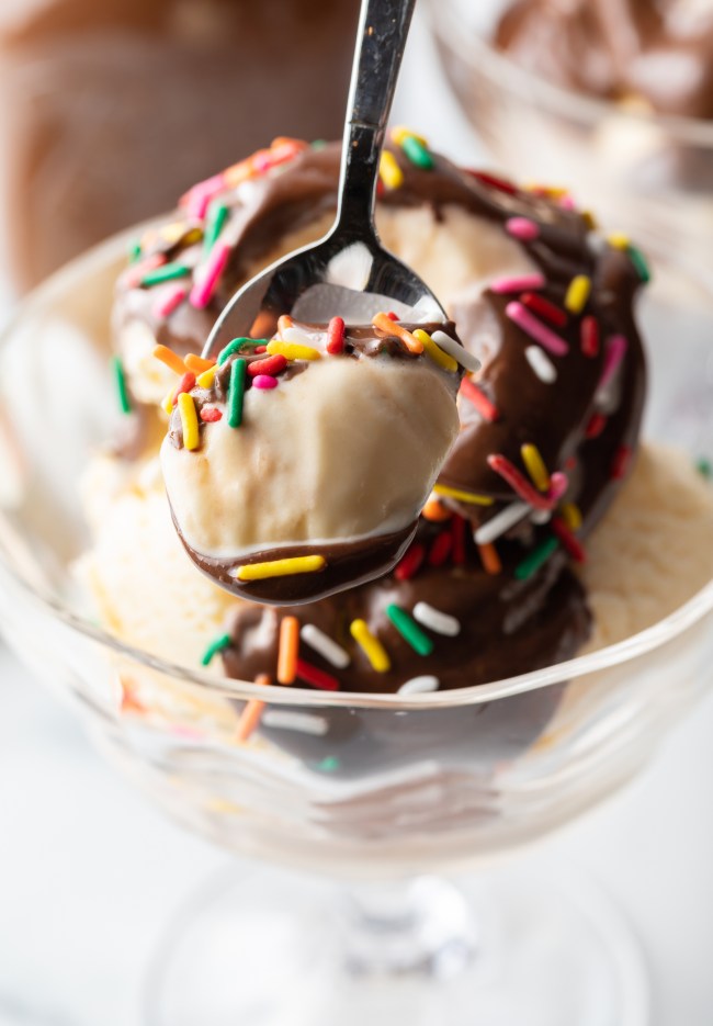 Metal spoon with scoop of vanilla ice cream, chocolate sauce, and sprinkles.