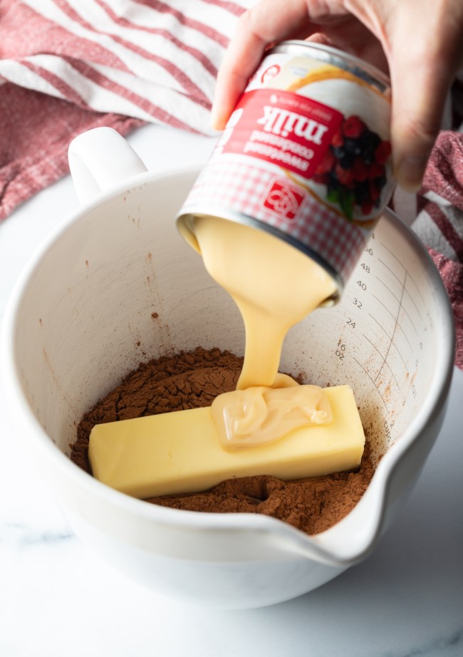 Hand adding condensed milk to a white bowl with cocoa powder and butter.