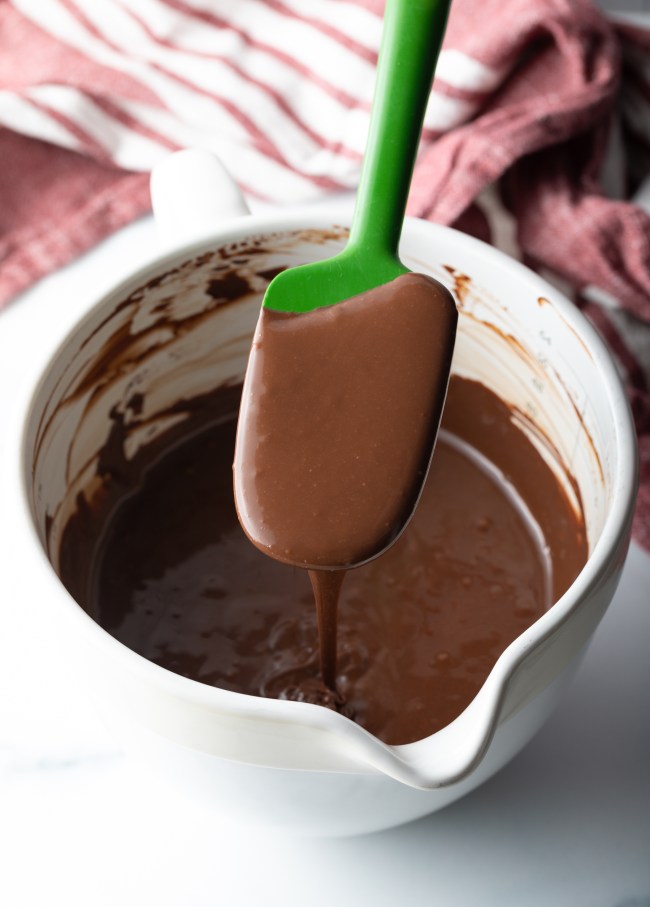 Green spatula held over a white bowl with homemade hot fudge sauce.