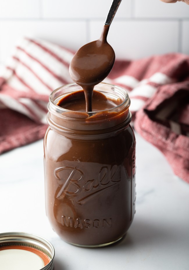 Metal spoon pouring homemade chocolate fudge sauce into a glass mason jar.