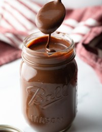 Metal spoon pouring homemade chocolate fudge sauce into a glass mason jar.