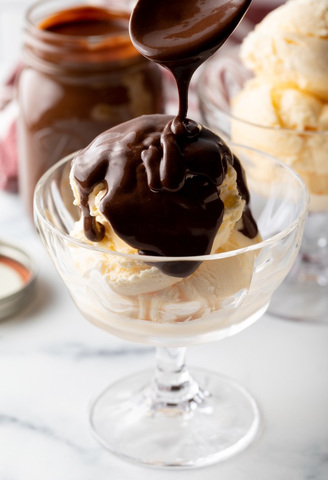 Spoon drizzling the homemade hotfudge sauce over a glass bowl of vanilla ice cream.