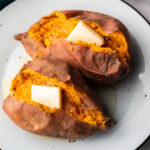 2 cooked sweet potatoes on a white plate, each is opened on top with a pat of butter inside.