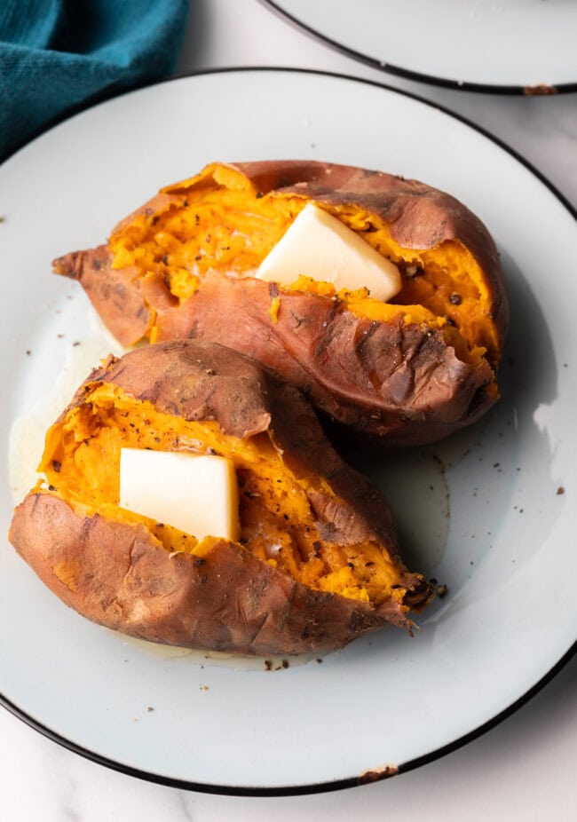 2 cooked sweet potatoes on a white plate, each is opened on top with a pat of butter inside.