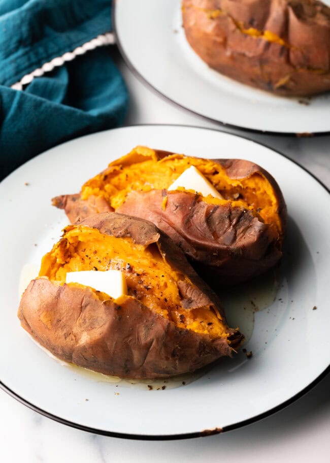 2 cooked potatoes on a white plate, each is opened on top with a pat of butter inside.