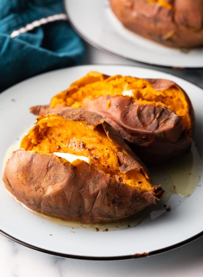 2 cooked sweet potatoes on a white plate, each is opened on top with a pat of butter inside.