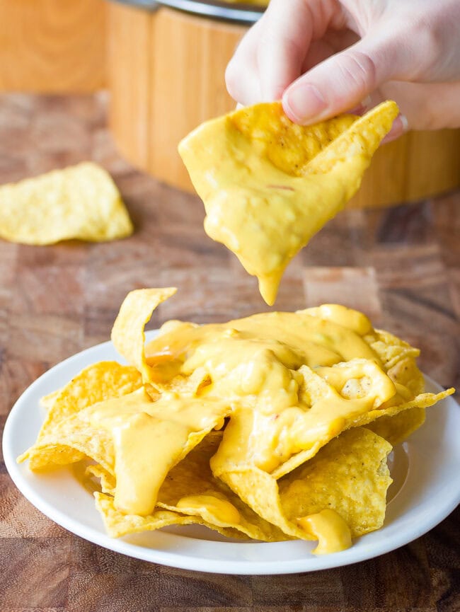 A hand taking a tortilla chip dipped in queso from a plate of queso-topped chips.