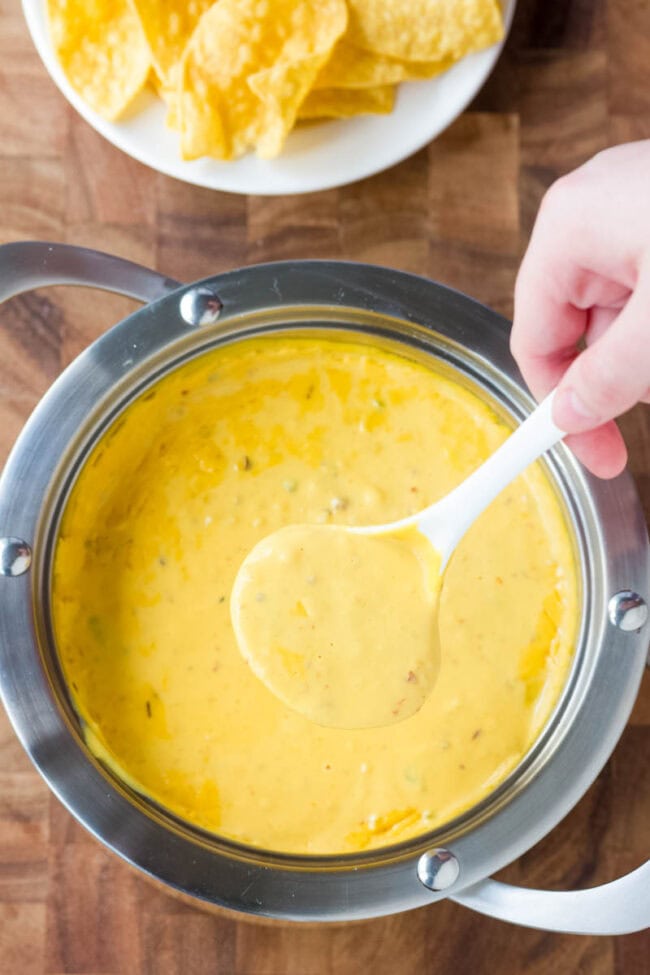 Hand holding a ladle of queso dip with the pot of more queso below it. 