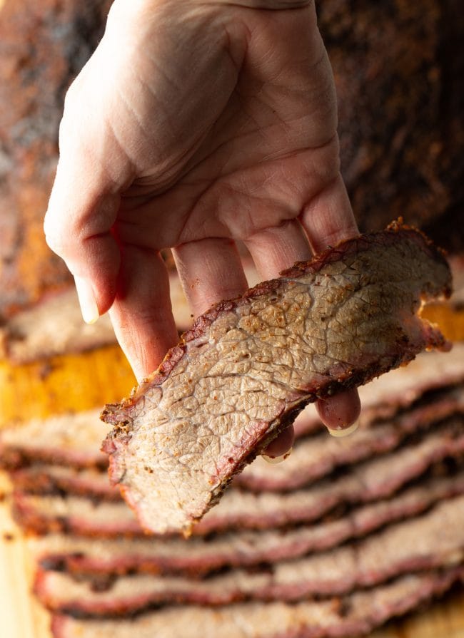 Hand holding a slice of brisket up close with more brisket in the background. 