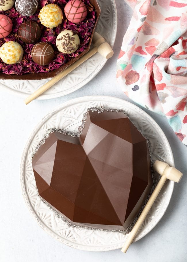 top view heart shaped chocolate box on a white plate with hammer, open bottom of the box with truffles in upper left corner