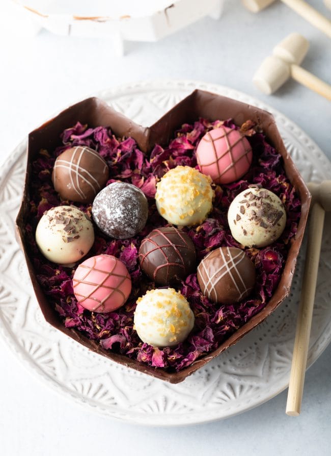 bottom half of breakable chocolate heart on a white plate filled with edible rose petals and various truffles