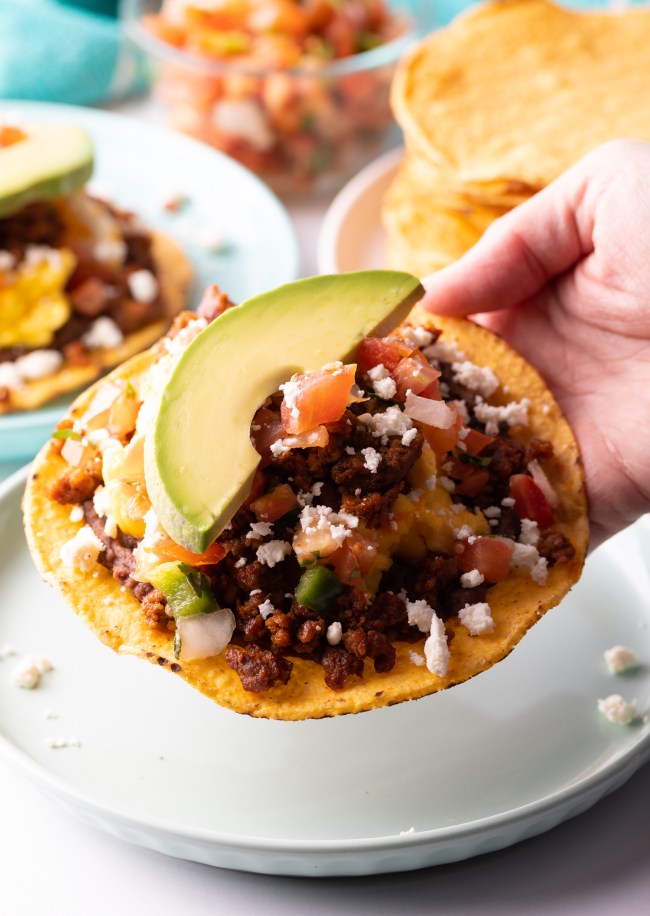 Hand holding tostada loaded with ingredients and slice of avocado to camera.