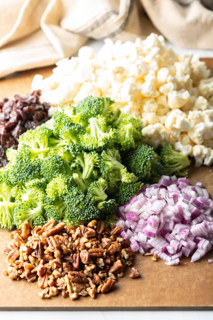 Cutting board with piles of fresh broccoli, cauliflower, chopped red onions, pecans, and cranberries.