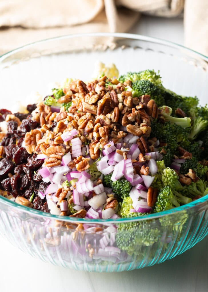 Large glass bowl with piles of fresh broccoli, cauliflower, chopped red onions, pecans, and cranberries.