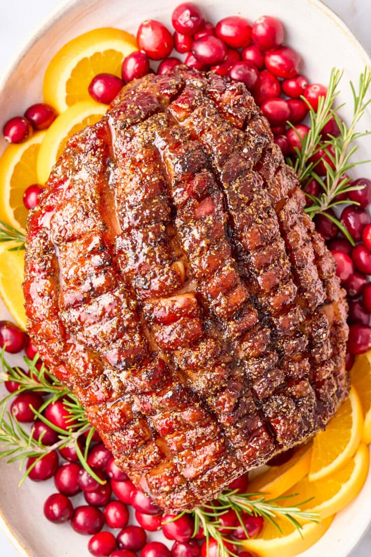 Overhead shot of brown sugar glazed ham on a serving platter with cranberries, oranges and rosemary. 