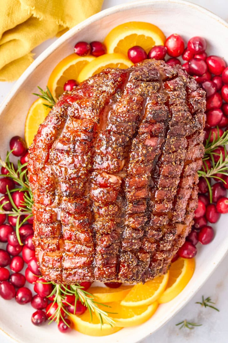 Baked Thanksgiving Ham with Brown Sugar Glaze on platter with oranges, cranberries, and herbs.