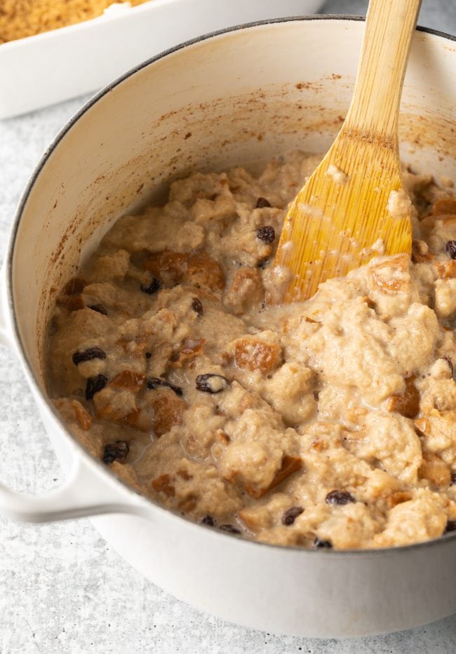 Pieces of bread being mushed by a wooden spatula in a white cooking pot, with raisins and spices.