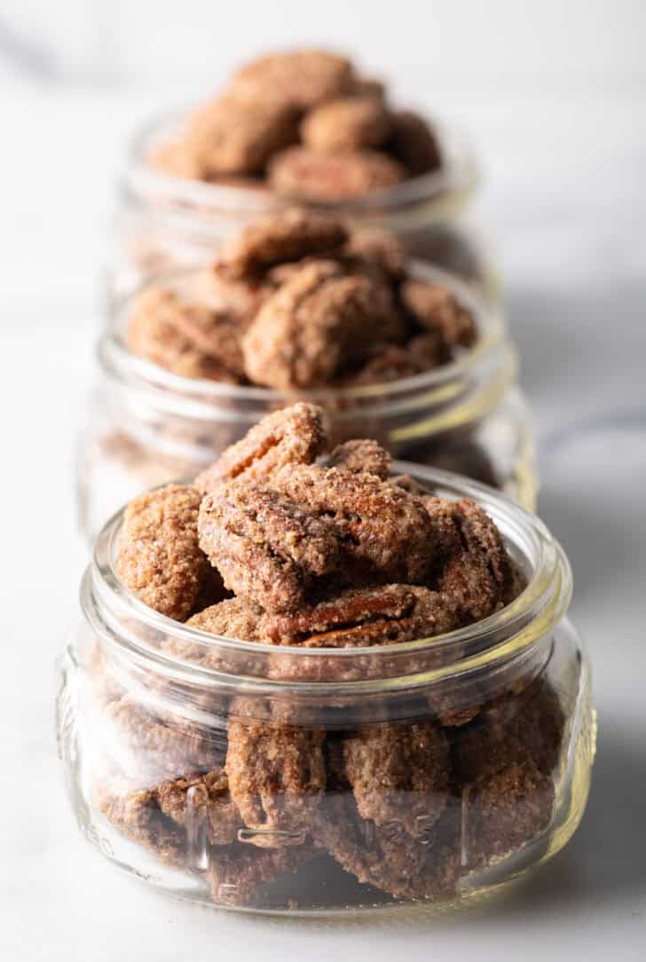 Close view of 3 small glass jars overflowing with candied pecans.