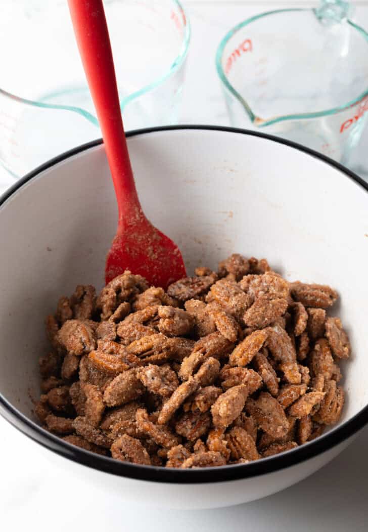 Red spatula stirring coated pecans in a large white mixing bowl.