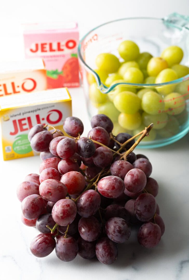 Recipe ingredients: a bunch of red grapes in the front view, in the background is a large glass measuring cup with green grapes. 3 boxes of Jello are to the side.