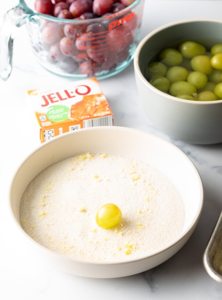 Shallow bowl with 1 green grape being rolled in a sugar and Jello powder mixture.