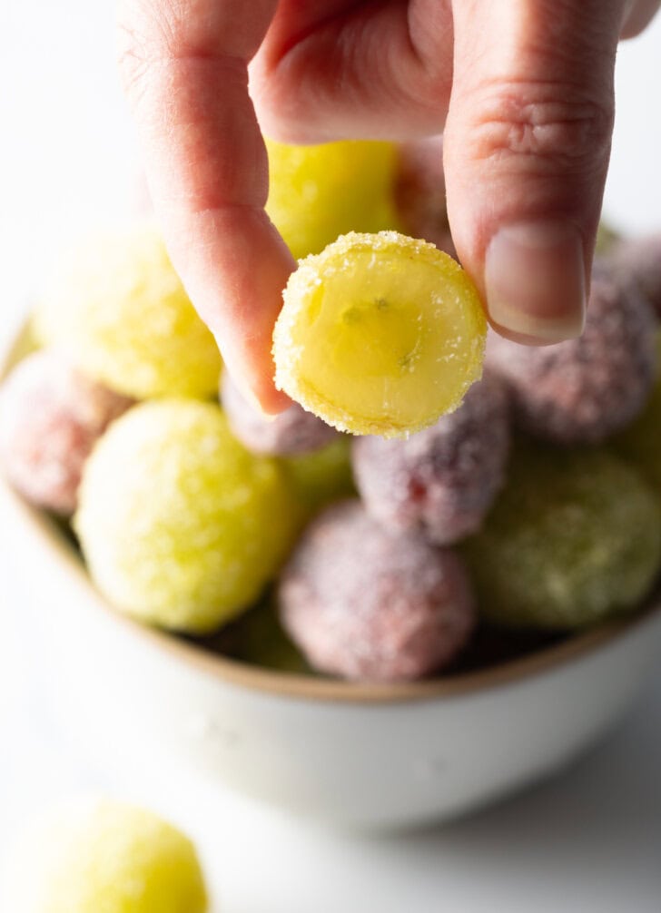 a bowl of green and purple candy grapes. A hand is holding one of the green grapes to camera. It has been bitten in half to show the inside.