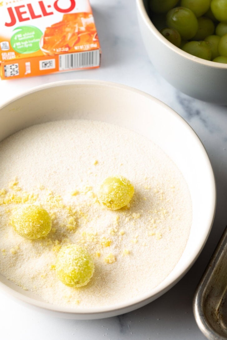 Shallow bowl with 3 green grapes being rolled in a sugar and Jello powder mixture.