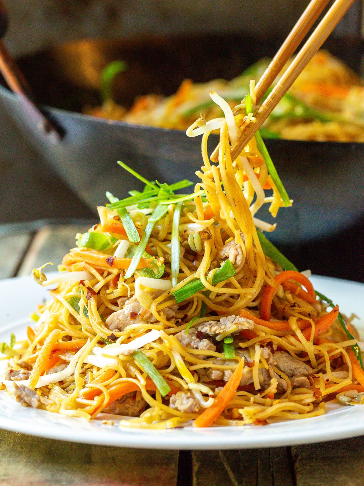 Cantonese Lo Mein piled on a white plate with a pair of chopsticks taking a bite. 