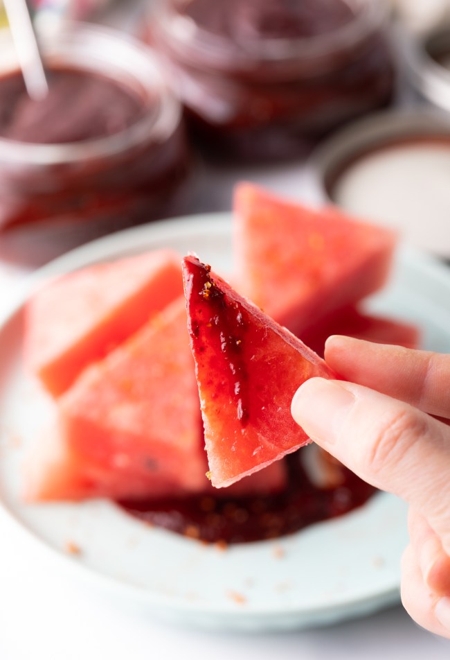 Hand holding small triangle of watermelon dipped in spicy sauce.