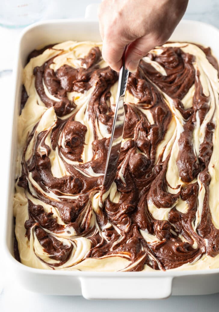 Hand with knife, swirling the raw cheesecake and brownie batter.