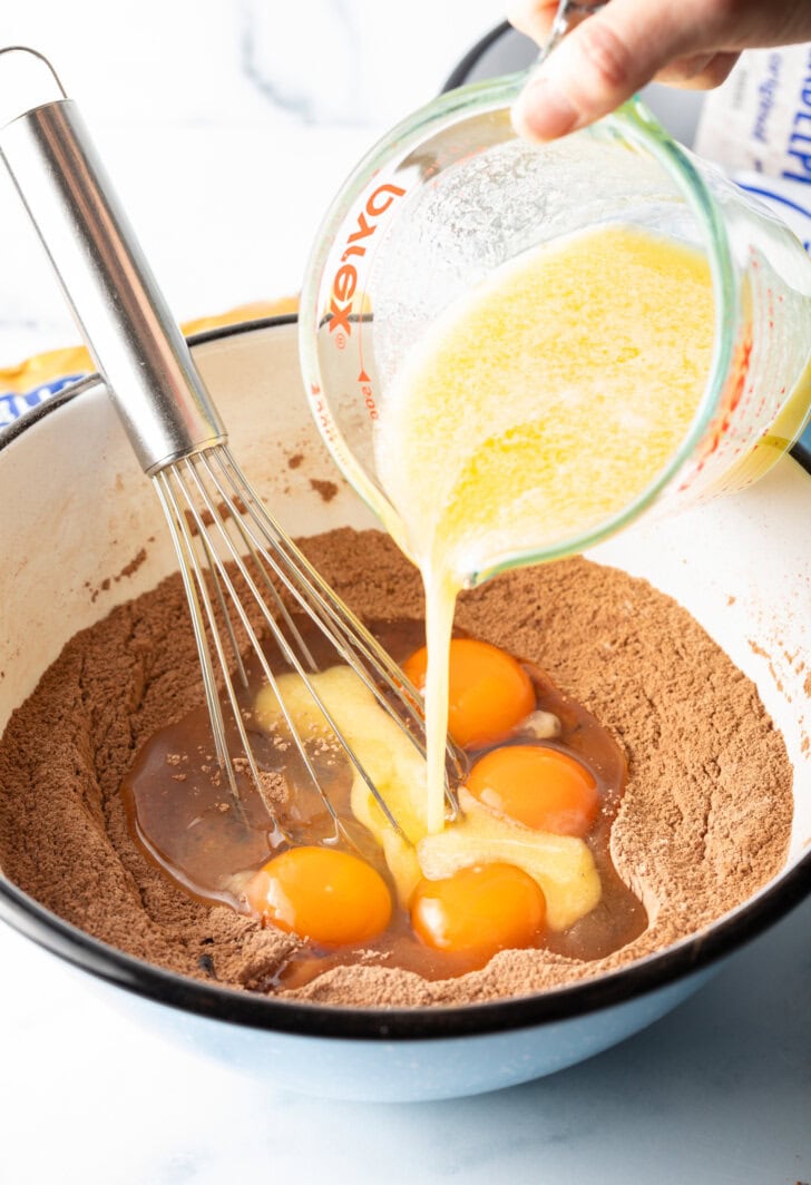 Adding butter from a glass measuring cup into a white bowl with dry ingredients and eggs.