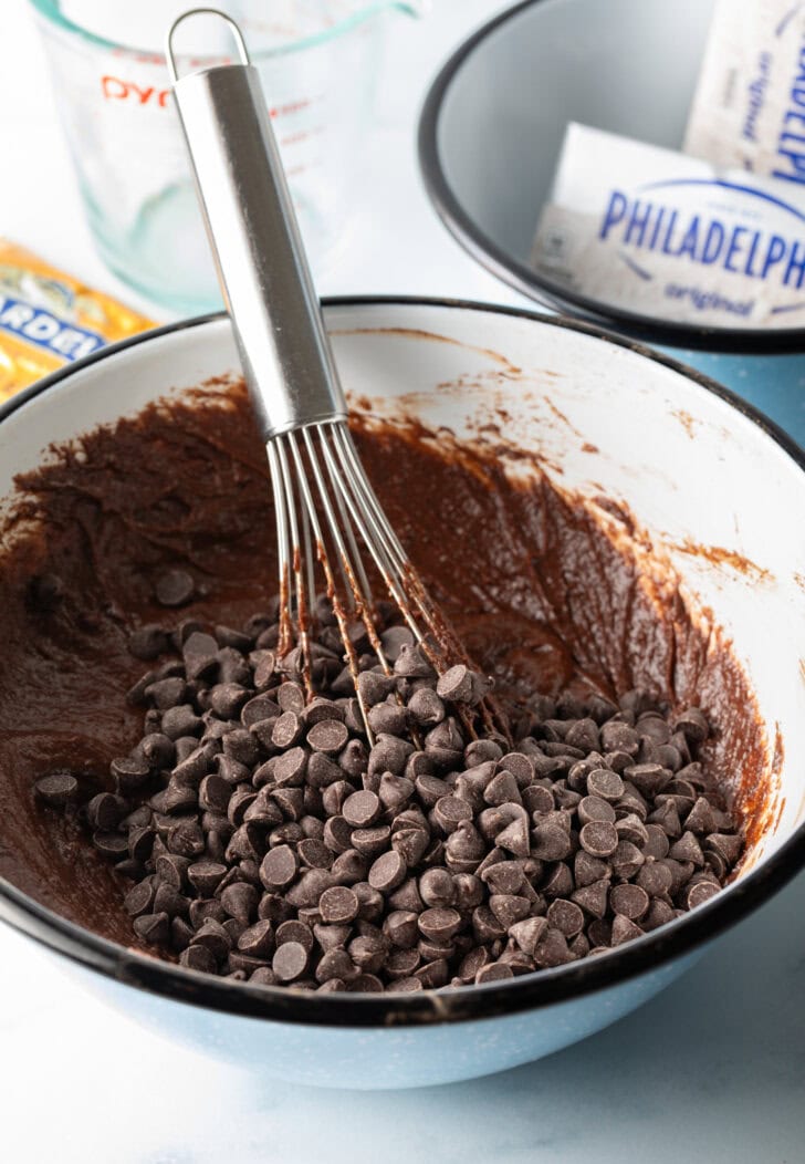 Whisking chocolate chips into the brownie batter in a white mixing bowl.