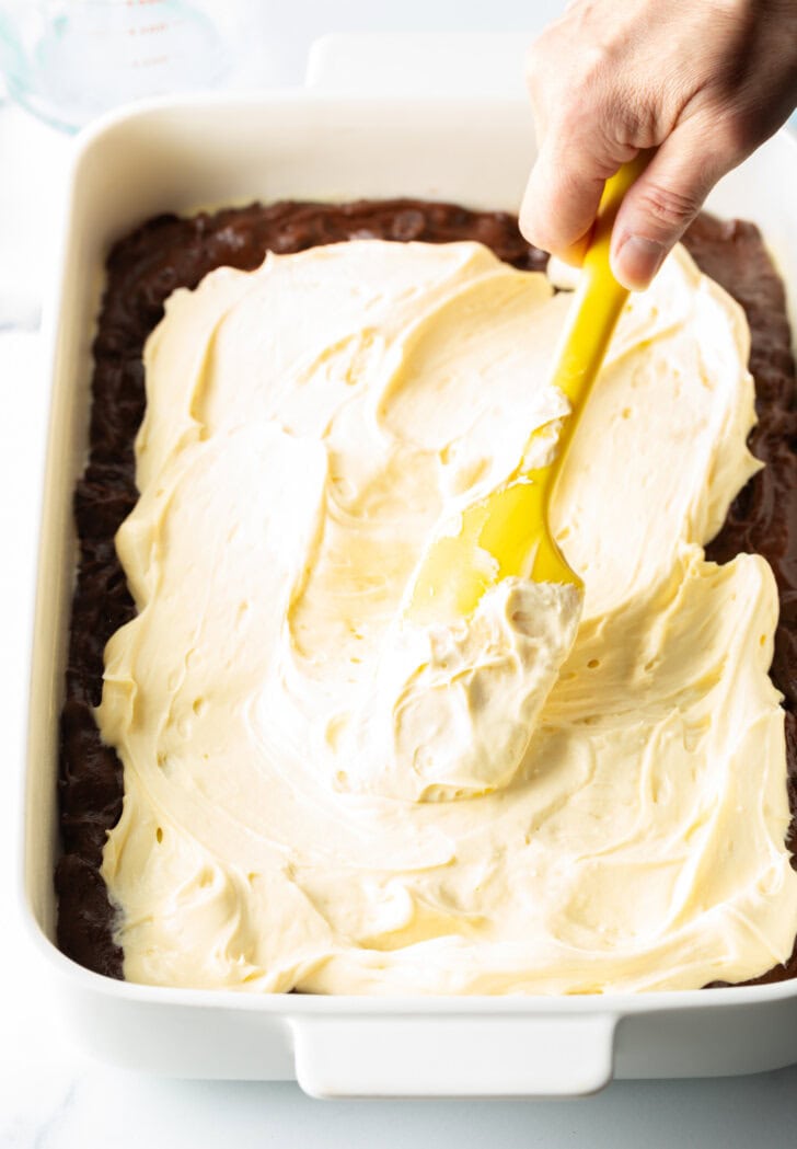 Hand using yellow spatula to spread cheesecake filling over the top of brownies.