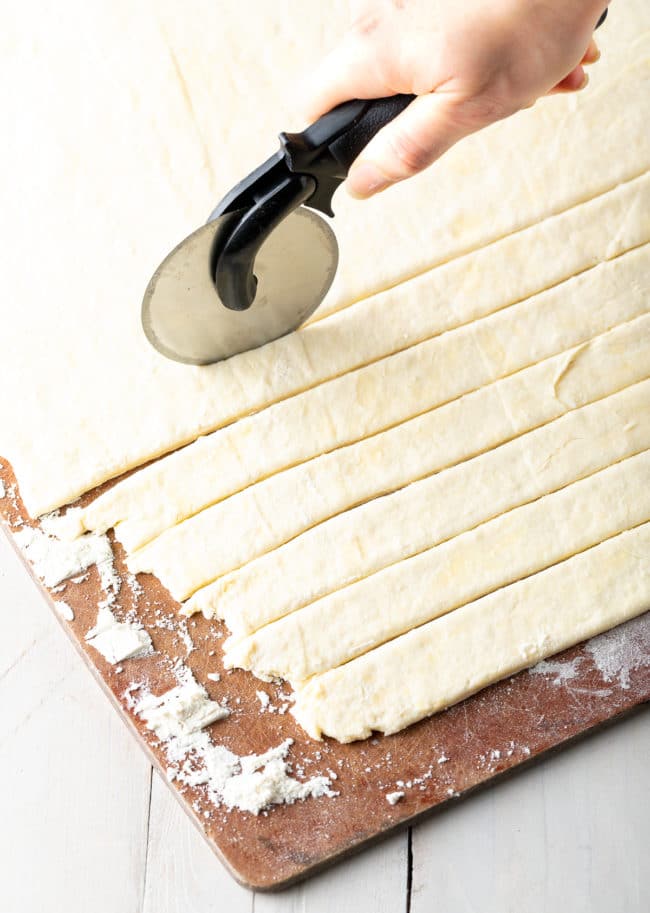 A pizza cutter cutting dumpling dough into long strips on a cutting board.